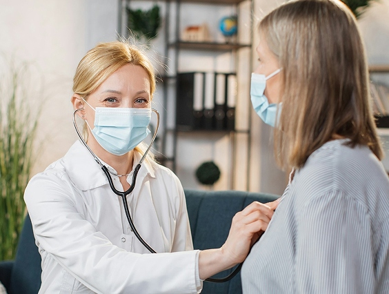 A healthcare professional wearing a mask uses a stethoscope to examine the chest of a patient, who is also masked, in a clinical setting.