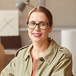 A woman with glasses and a light brown jacket sits indoors, smiling at the camera. There are office supplies and a lamp in the blurred background.