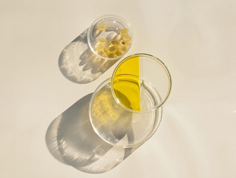 A glass dish with yellow liquid and a small round container holding yellow capsules on a light surface, casting shadows.